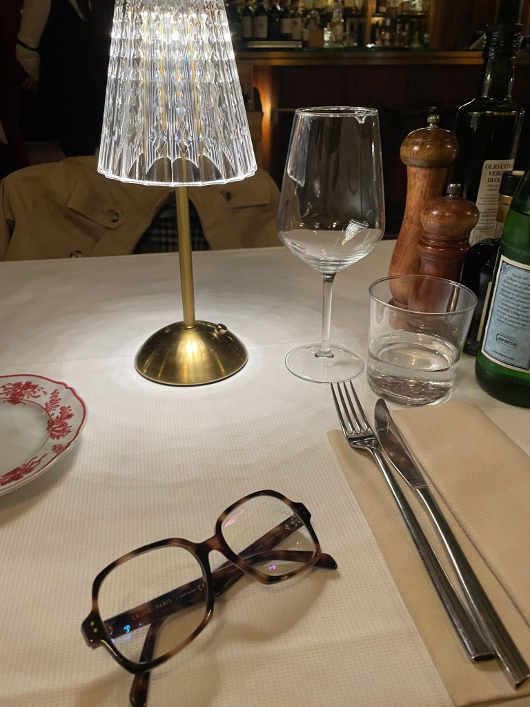 Reading glasses on a restaurant table laid for one person in dimmed evening light.